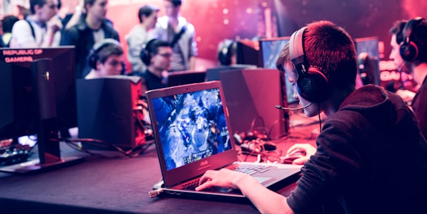 Several people are seated and focused on computer screens, wearing headsets, in an environment that appears to be a gaming tournament or esports event. The room is dimly lit with neon lighting, highlighting screens and creating an intense atmosphere.