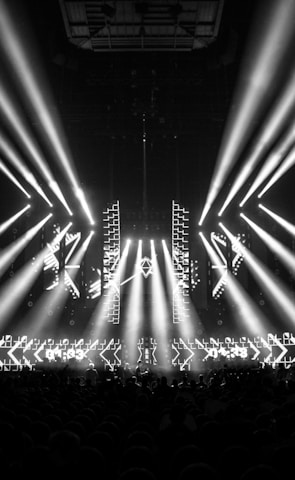 A black and white image captures a dramatic indoor scene featuring a large stage setup with intense beams of light radiating from the center. Two large screens are prominently displaying the text '01:33 ESL One Cologne 2017'. The audience can be seen seated in the dark foreground, while angular patterns can be spotted on the stage.