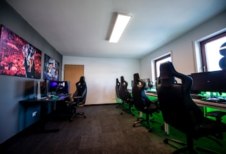 A well-lit gaming room features multiple gaming stations with high-backed chairs and monitors. The room has a minimalist setup with green and black color tones. Large posters adorn the walls, and the room appears clean and organized.