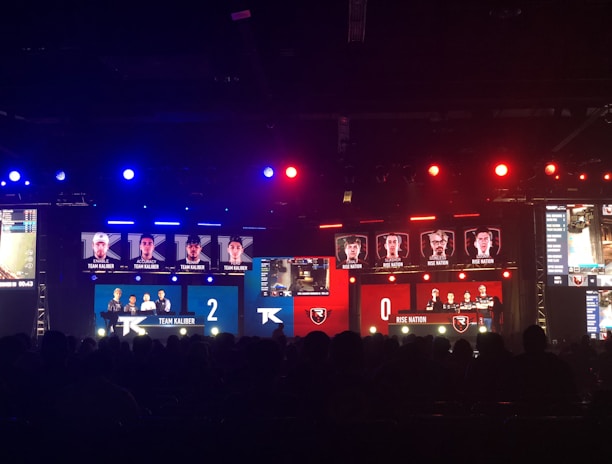 An esports event with a large screen displaying two teams, Team Kaliber and Rise Nation, competing against each other. The screen shows profiles of team members and scores, with Team Kaliber leading 2-0. The venue is dimly lit with blue and red lighting, and an audience can be seen watching.