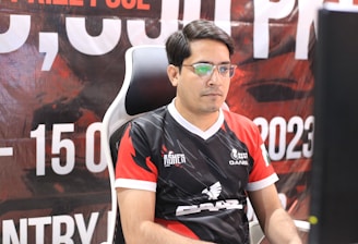 A man with glasses is sitting in a gaming chair, focused on a monitor in front of him. He is wearing a black and red esports jersey with various logos. The background features a large banner with text and numbers in bold fonts.