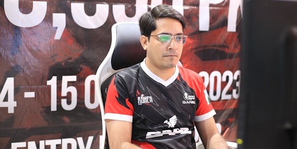 A man with glasses is sitting in a gaming chair, focused on a monitor in front of him. He is wearing a black and red esports jersey with various logos. The background features a large banner with text and numbers in bold fonts.