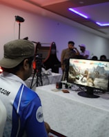 A group of people is gathered in a room watching and participating in a video game competition. Players and spectators are focused on the monitors displaying a fighting game. The setting includes tables with equipment and chairs arranged around the venue, with a noticeable intensity in the participants’ engagement.