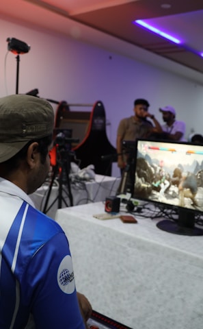 A group of people is gathered in a room watching and participating in a video game competition. Players and spectators are focused on the monitors displaying a fighting game. The setting includes tables with equipment and chairs arranged around the venue, with a noticeable intensity in the participants’ engagement.